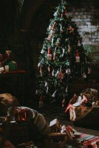 A decorated Christmas tree stands indoors with gifts, toys, and wrapped presents on the floor around it in a dimly lit room.