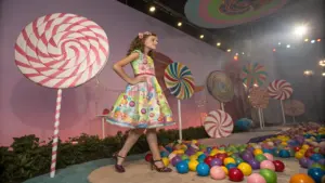 A young girl in a colorful candy-themed dress walks on a set decorated with oversized lollipops and multicolored plastic balls.