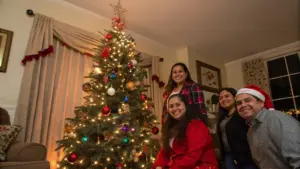 Four people pose and smile beside a decorated Christmas tree with lights and ornaments in a living room. One person wears a Santa hat.
