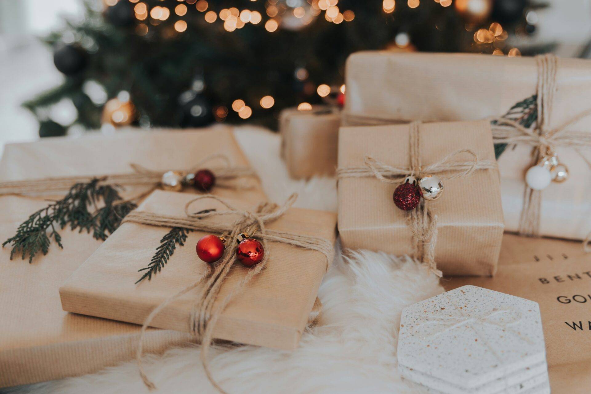 Several gifts wrapped in brown paper and twine decorated with small ornaments and pine sprigs are placed on a white surface in front of a lit Christmas tree.