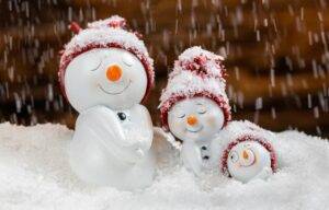 Three small snowman figurines with red hats sit in snow as artificial snow falls around them. The background is blurred and brown.