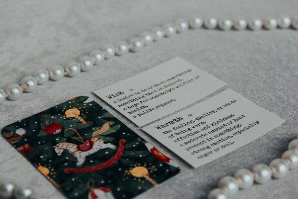 Three cards lie on a gray surface with a string of pearls; two cards have dictionary-style definitions, and one card has a holiday-themed rocking horse design.