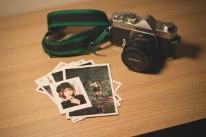 A Konica film camera with a green strap sits on a wooden surface next to several printed photographs, including portraits and an outdoor scene.