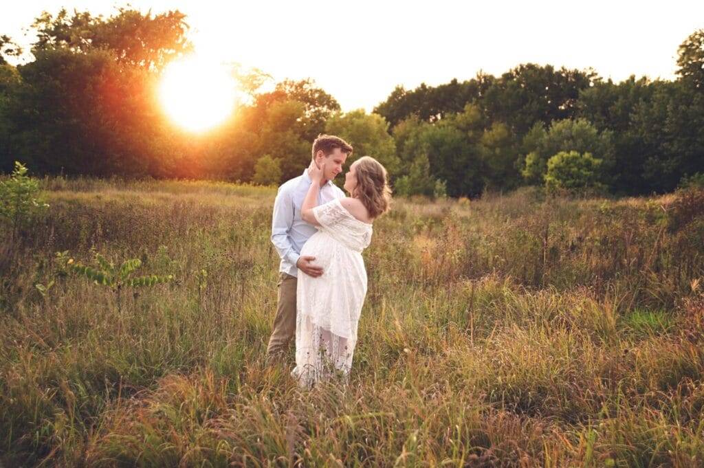 A couple stands in a grassy field at sunset, with the woman in a white dress holding her pregnant belly and the man gently touching her face.