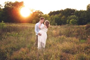 A couple stands in a grassy field at sunset, with the woman in a white dress holding her pregnant belly and the man gently touching her face.