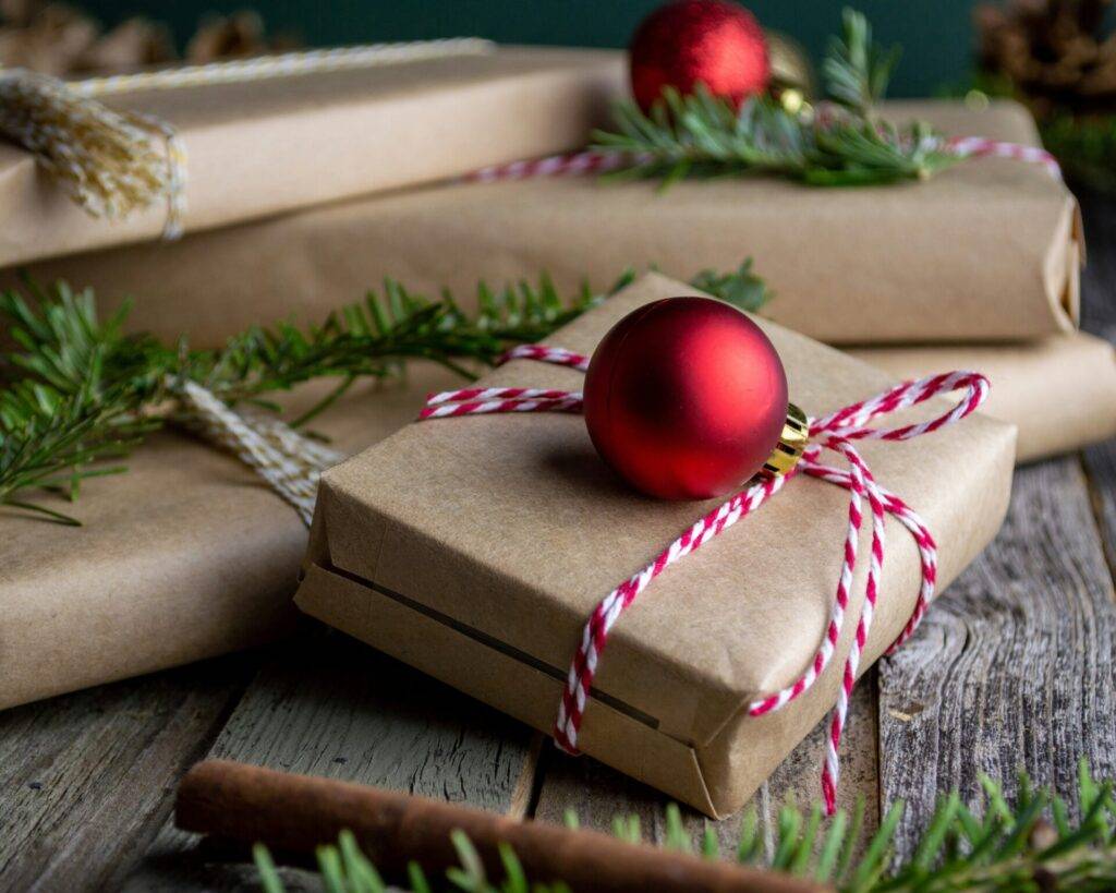 Several brown paper-wrapped gifts with red and white string, decorated with pine branches and red Christmas ornaments, are arranged on a rustic wooden surface.