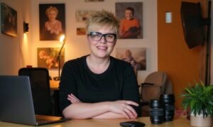 A person with short blond hair and glasses sits at a desk with a laptop, camera lenses, and a plant, with portrait photos displayed on the wall behind them.