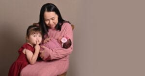 A woman in a pink dress holds a swaddled baby while a young girl in a red dress sits beside them, making a shushing gesture.