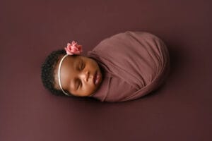 A newborn baby wrapped in a mauve blanket lies on a matching background, wearing a pink flower headband and sleeping peacefully.