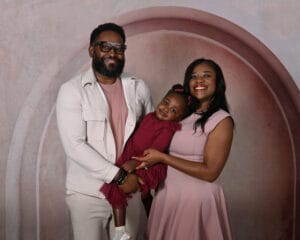 A man and woman stand together smiling, holding a young girl in a maroon dress. All three are dressed in coordinating pink and maroon tones against a pink and mauve arched backdrop.