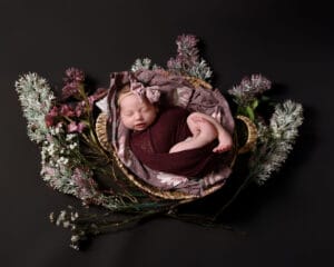 A newborn baby, wrapped in a burgundy cloth with a matching headband, sleeps in a basket surrounded by artificial flowers on a dark background.