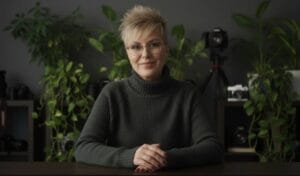 A person with short blond hair and glasses sits at a table, wearing a dark sweater, with cameras and green plants in the background.