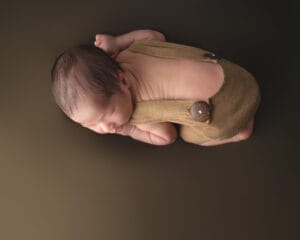 A newborn baby in a brown outfit is lying on their side on a plain brown background, with knees tucked under the chest and arms drawn in.