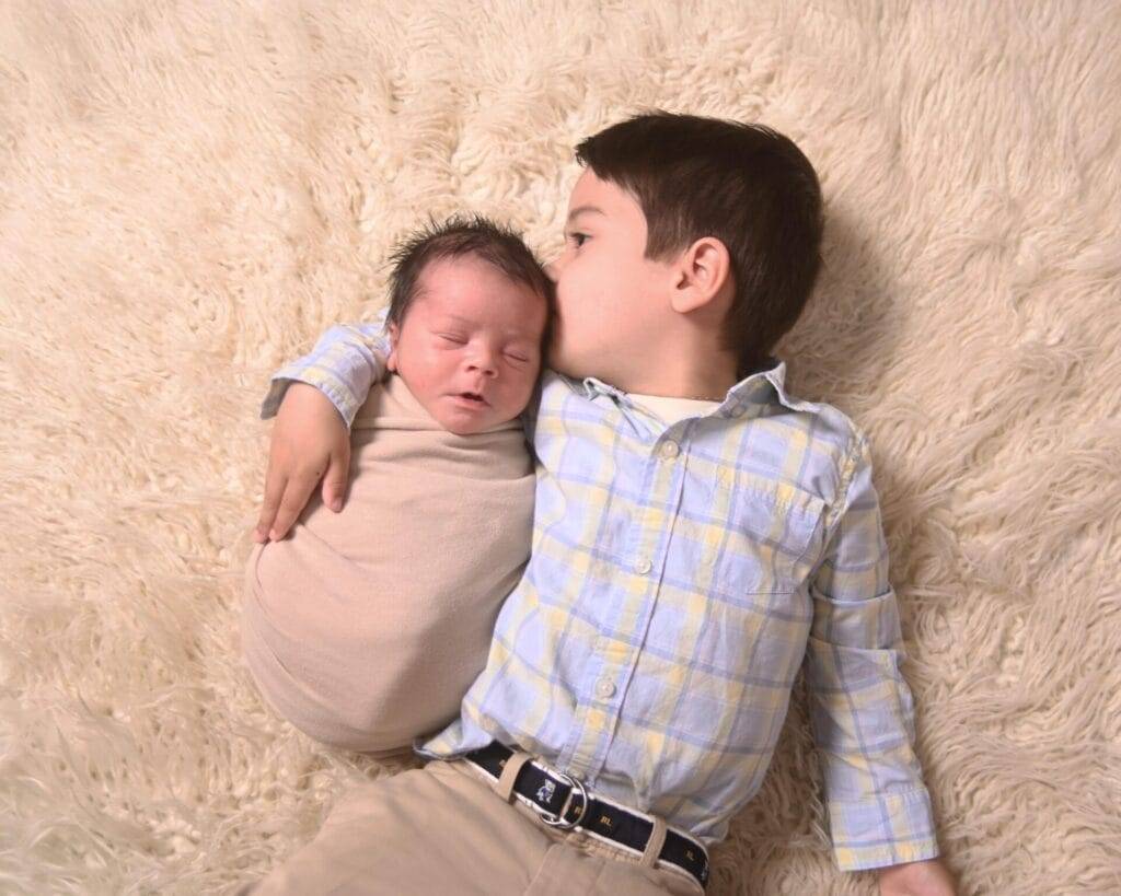 A young boy lies on a soft beige rug, gently holding and kissing a swaddled newborn baby beside him.