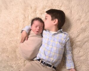 A young boy lies on a soft beige rug, gently holding and kissing a swaddled newborn baby beside him.
