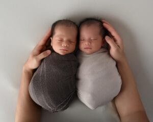Two newborn babies swaddled in brown and light gray blankets lie side by side, cradled by an adult’s hands, against a plain background.