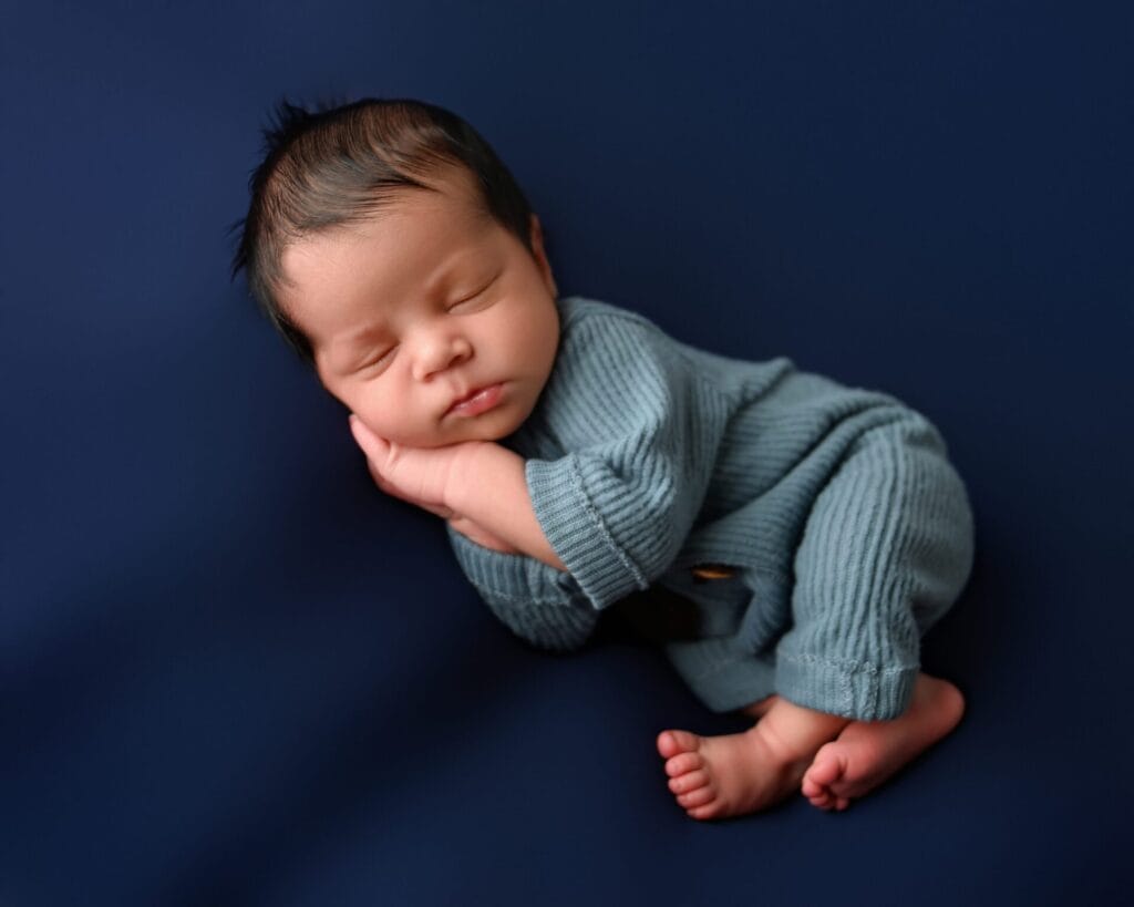 A newborn baby in a blue outfit sleeps on a dark blue background with one hand resting under their cheek and legs curled up.