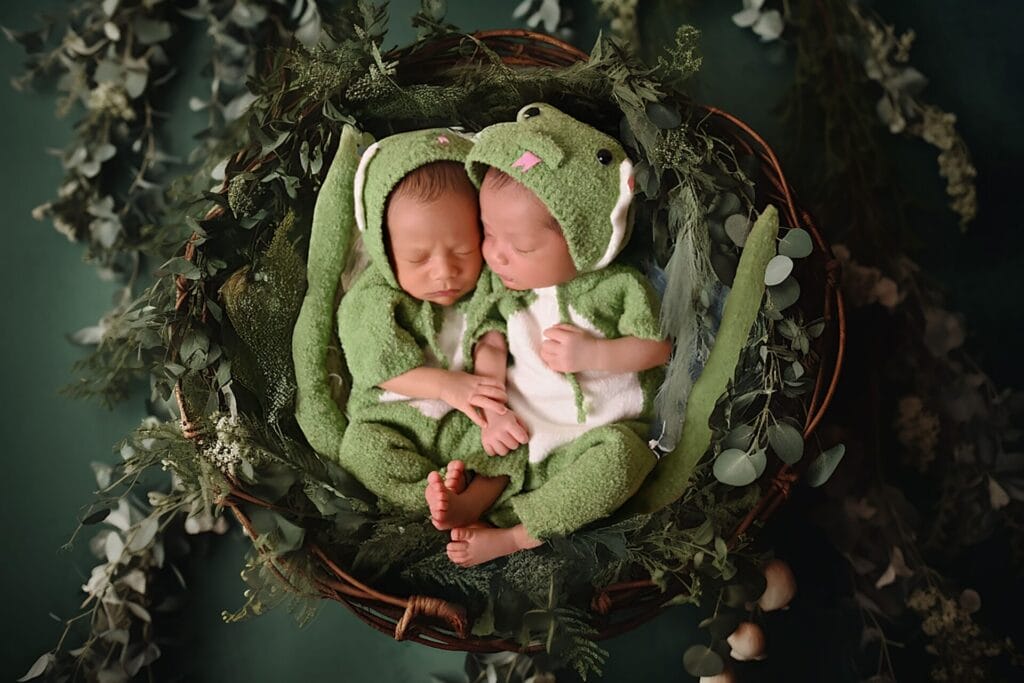 Two sleeping babies dressed in green frog costumes lie side by side in a basket surrounded by greenery.