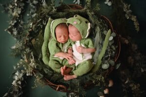 Two sleeping babies dressed in green frog costumes lie side by side in a basket surrounded by greenery.