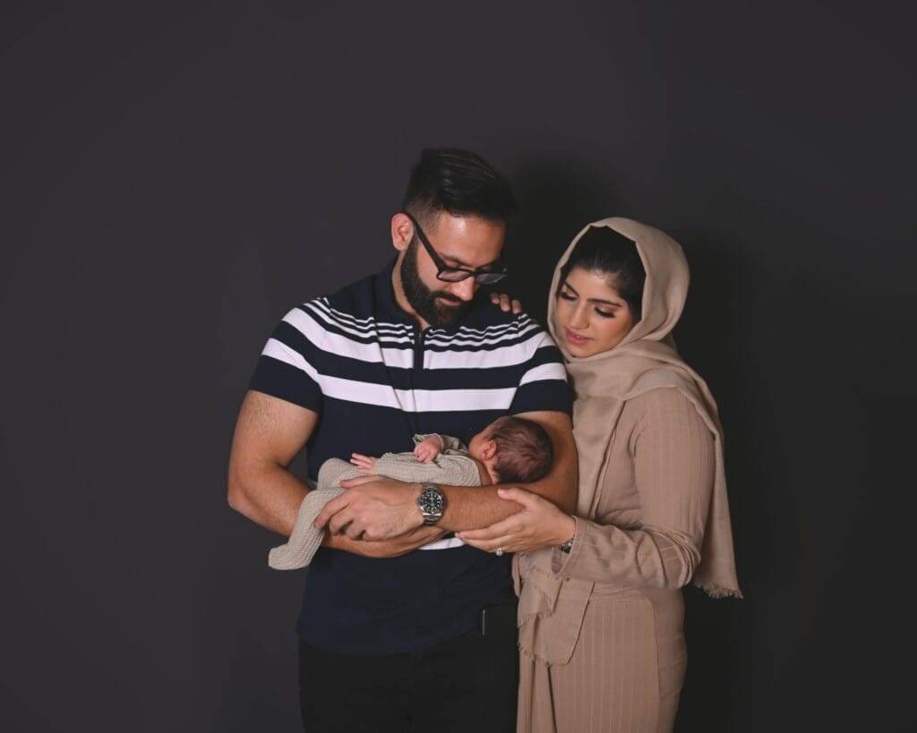 A man and woman stand together against a dark background, looking down at a baby the man is holding in his arms.