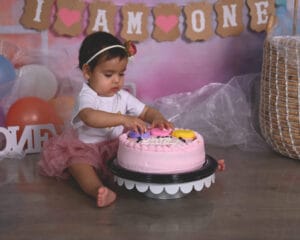 A baby wearing a pink skirt and white shirt touches a pink cake while sitting on the floor in front of a “I am one” birthday banner.