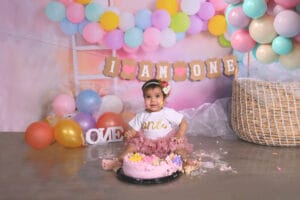 A baby in a "one" t-shirt and pink tutu sits in front of a smashed pink cake, surrounded by colorful balloons and a "I AM ONE" banner, celebrating a first birthday.