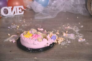 A smashed pink cake with colorful frosting sits on the floor, surrounded by crumbs and mess, with balloons and a "ONE" sign in the background.