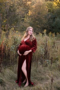 A pregnant woman in a long, red velvet dress stands barefoot in a grassy, wooded area during daylight.