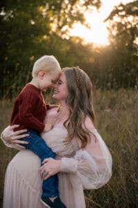 A woman in a light dress holds a young child in a red sweater as they touch foreheads and smile in a grassy field at sunset.