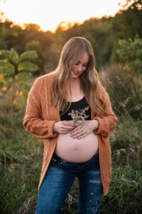 A pregnant woman stands outdoors at sunset, wearing a brown cardigan and jeans, holding dried flowers and looking down at her exposed belly.