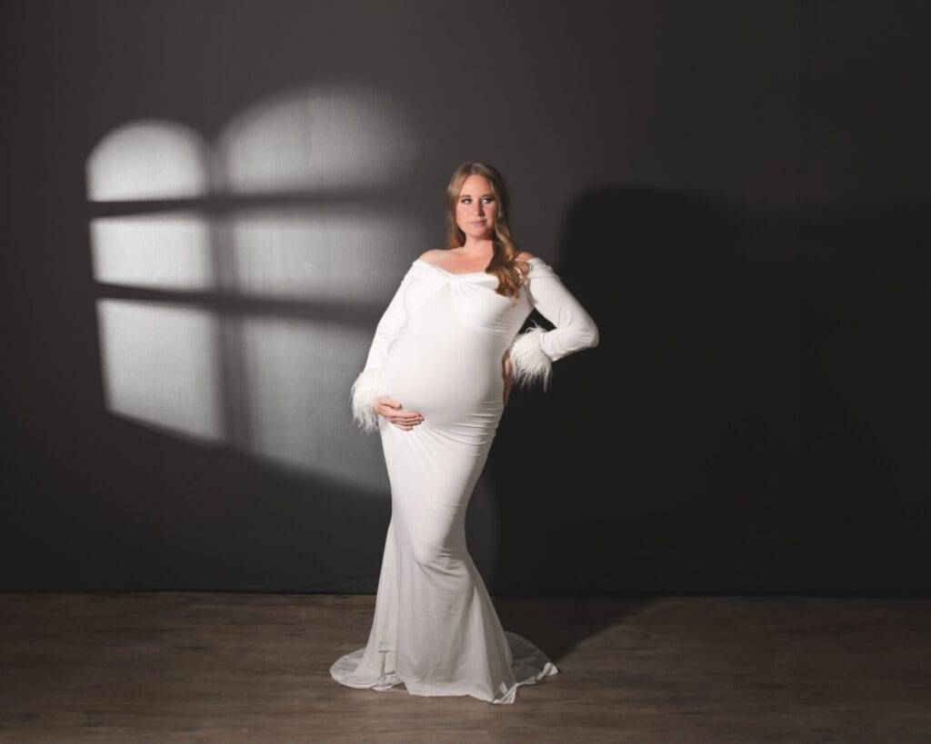 A pregnant woman in a long white dress poses against a dark wall with a window-shaped light pattern, one hand on her hip and the other on her belly.