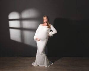 A pregnant woman in a long white dress poses against a dark wall with a window-shaped light pattern, one hand on her hip and the other on her belly.