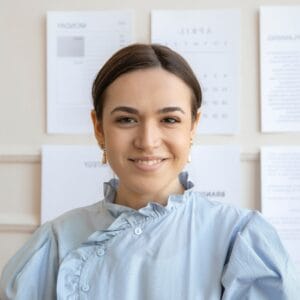 A woman in a light blue blouse is smiling at the camera, standing in front of a wall with several sheets of paper and documents displayed.