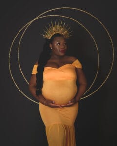 A pregnant woman in a yellow off-shoulder dress poses against a dark background, wearing a gold spike headpiece with gold rings behind her.