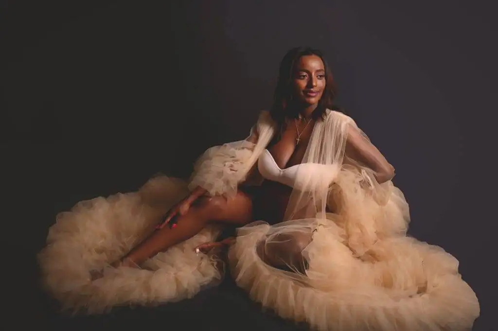 A woman in a white bra and sheer, ruffled beige robe sits on the floor against a dark background, looking slightly to the side and smiling.