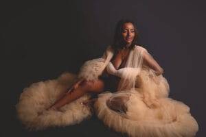A woman in a white bra and sheer, ruffled beige robe sits on the floor against a dark background, looking slightly to the side and smiling.