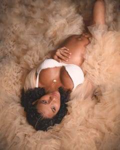 A pregnant woman in a white bra and beige tulle skirt lies on her back, looking up at the camera with one hand on her belly.