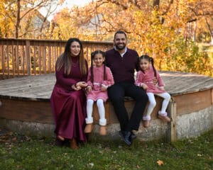 A family of four sits together on a wooden platform outdoors in autumn, all wearing coordinated burgundy and pink outfits and smiling at the camera.
