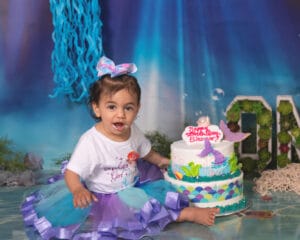 A toddler in a purple and blue tutu sits in front of an ocean-themed birthday cake with a “Happy Birthday Eleanor” topper in an under-the-sea decorated setting.