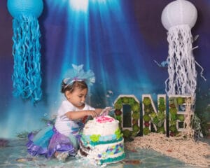 A toddler in a blue and purple tutu sits in front of a decorated cake at an under-the-sea themed first birthday party with large "ONE" letters and jellyfish decorations in the background.