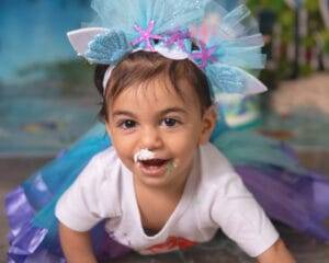 A young child in a blue and purple tutu and a sea-themed headband crawls on the floor with cake or frosting on their nose and mouth.