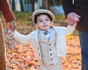 A young child in a beige suit, tie, and flat cap holds hands with two adults while standing on fallen autumn leaves.