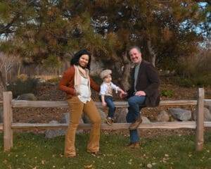 Three people, two adults and a child, posing outdoors on a wooden fence in front of trees and rocks, dressed in fall clothing.