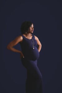 A pregnant woman in a sleeveless, dark blue dress stands against a dark background, looking to the side with one hand on her hip.