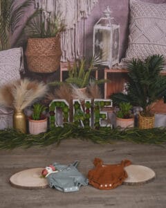 Two baby outfits, one green and one brown, are laid on wood slices in front of a decorated "ONE" sign, plants, and rustic decor against a macramé backdrop.