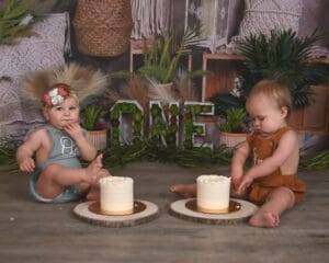 Two babies sit on the floor in front of small cakes, with "ONE" displayed behind them, surrounded by plants and decorations.