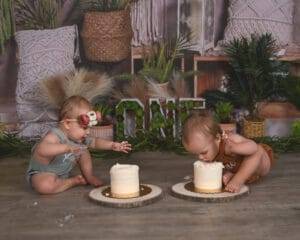 Two babies in front of greenery and “ONE” sign celebrate a first birthday; one baby leans to eat cake face-first, the other reaches toward their own cake.