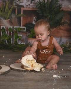 A baby wearing a brown romper with "one" on it sits on the floor, covered in cake and frosting, with a smashed cake in front of them.
