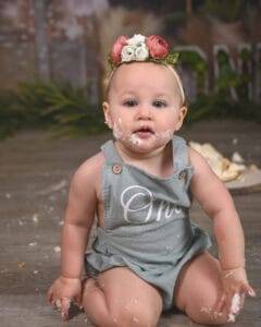 A baby wearing a gray romper and floral headband sits on the floor with cake and frosting smeared on their face and hands.