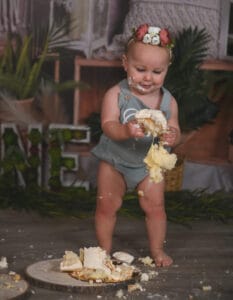 A baby in a decorative floral headband and grey romper stands, holding and smashing a piece of cake, with cake and crumbs scattered on the floor.